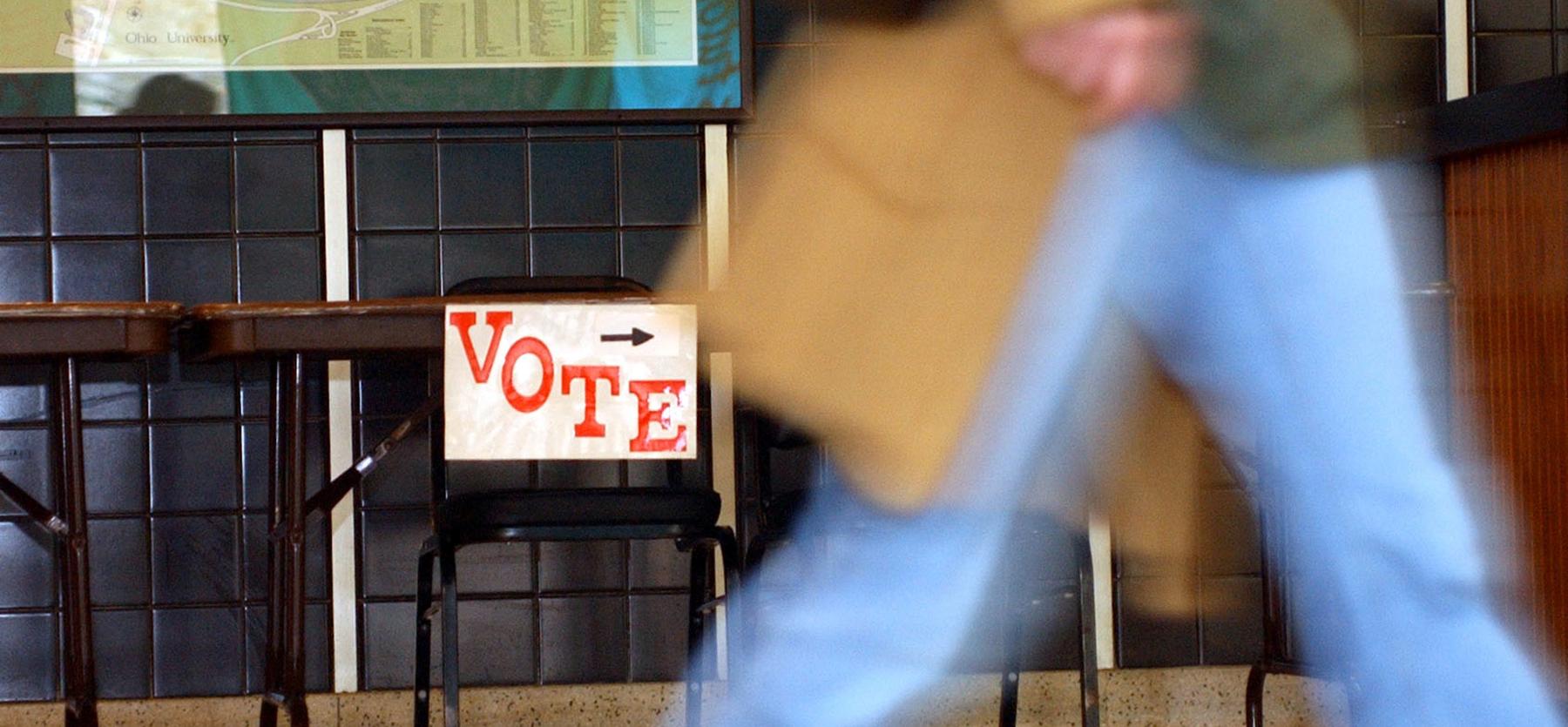 Out-of-focus person walks past a sign on a table that says "vote"