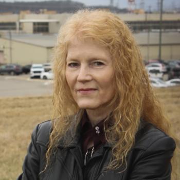 Headshot of smiling woman with long curly strawberry blonde, curly hair wearing a black coat infront of an industrial site