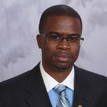 Professional headshot of Floun'Say Caver, Black man with glasses wearing a dark suit, blue dress shirt with white color and tie. 