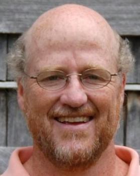 Headshot of Sam Crowl. Man smiling at camera wearing wire framed glasses. He has a short red-tinted beard, short graying hair over his ears and is wearing a coral shirt. He is standing infront of a background of weathered, graying wood.