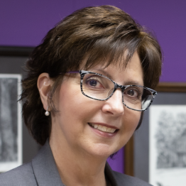 White woman smiles with short brown hair, glasses and a gray suit coat infront of a purple wall with framed art