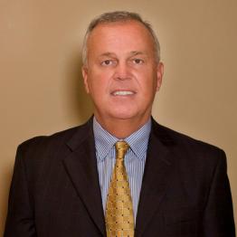 Bret Adams Headshot: Caucasian man wearing a dark suit jacket, mustard tie and blue dress shirt. He had short gray hair and is standing infront of a tan wall. 