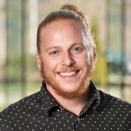 Headshot of Shawn Carvin. Caucasian man with red hair and short beard. He is wearing a black and white dress shirt with a patterned background.