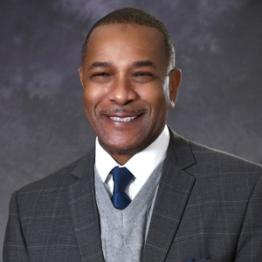 Doug Franklin Headshot. African American Man wearing a charcoal suit jacket, gray sweater with navy tie. 