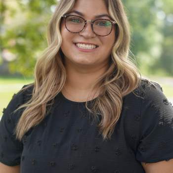 Headshot of Alyssa Manning. Woman smiles at the camera, she is wearing a navy blouse and dark rimmed glasses. Her blonde and brown hair hangs past her shoulders and her backdrop is an outdoor scene of trees and grass. 