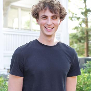 A headshot of Maxwell Burton. A man smiles at the camera and is wearing a black t-shirt. He has curly brown hair and the background of the image is of a white porch and landscaping.